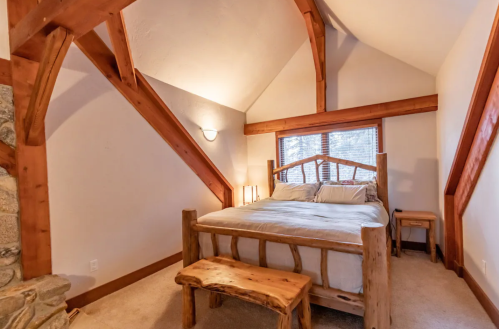 Cozy bedroom with a wooden bed, bench, and sloped ceiling, featuring large windows and warm lighting.